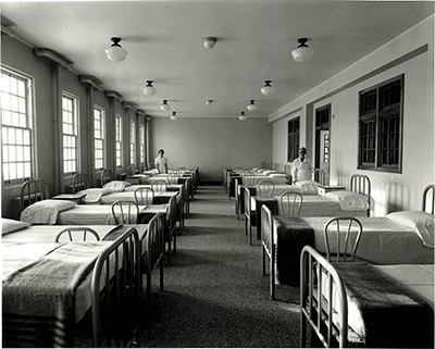 institutional ward with several empty beds and two stern-looking nurses